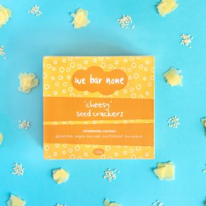 We Bar None Cheesy seed crackers. A box lying on a blue background surrounded by little piles of cheese and sesame seeds.