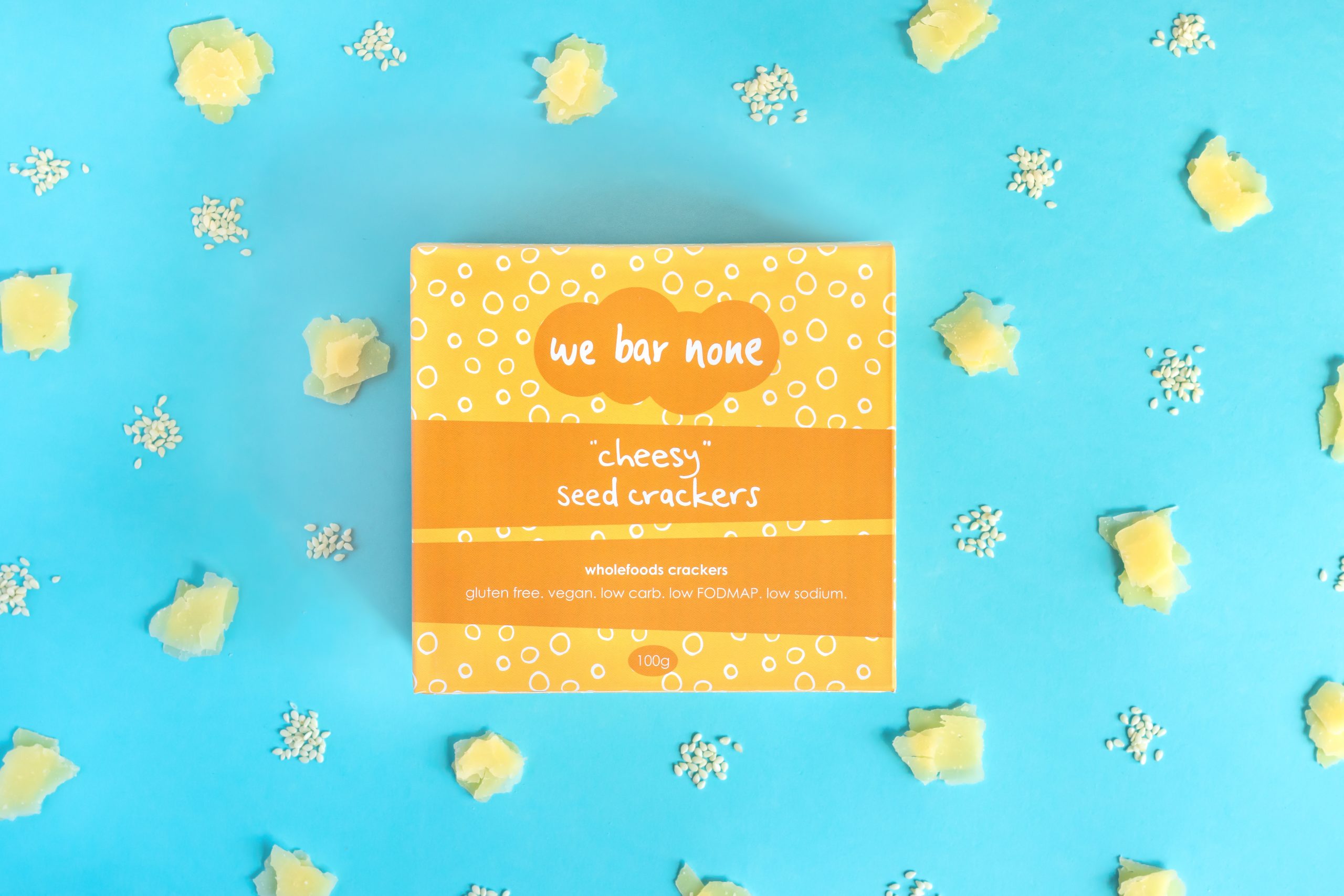 We Bar None Cheesy seed crackers. A box lying on a blue background surrounded by little piles of cheese and sesame seeds.
